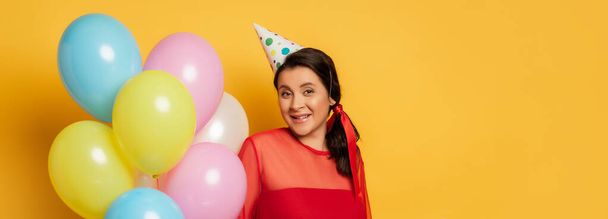 horizontal image of young woman in party cap holding multicolored festive balloons on yellow - Fotografie, Obrázek