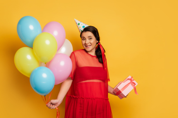 беременная женщина в кепке с подарочной коробкой и разноцветными праздничными воздушными шарами на желтом - Фото, изображение