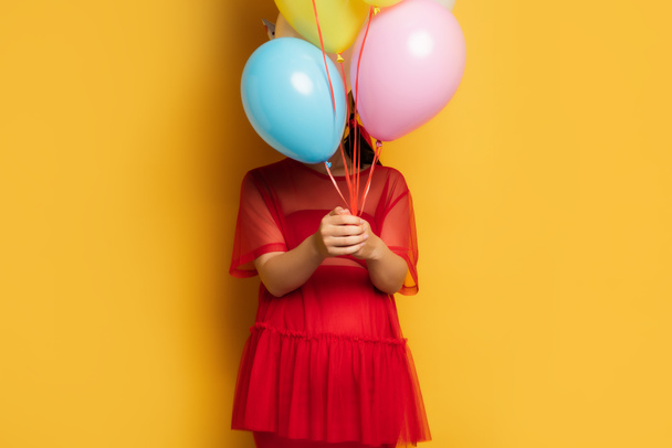 young pregnant woman in red outfit obscuring face with colorful festive balloons on yellow - Фото, изображение