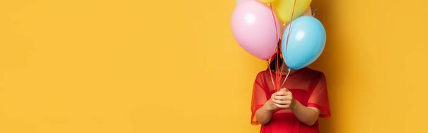 horizontal image of pregnant woman in red tunic obscuring face with multicolored festive balloons on yellow - Foto, Imagen