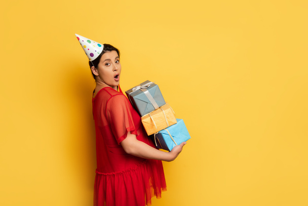 shocked pregnant woman in party car holding stack of gifts while looking at camera on yellow - Foto, immagini