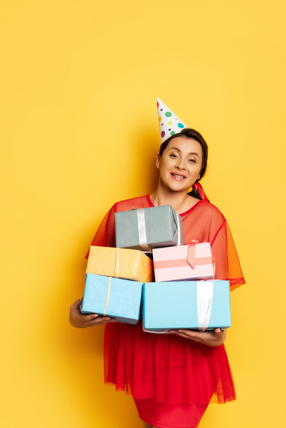 young pregnant woman in party cap holding stack of gift boxes on yellow - Foto, imagen