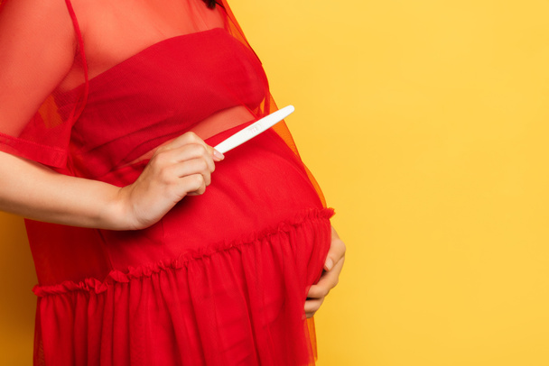 abgeschnittene Ansicht einer schwangeren Frau in rotem Outfit, die einen Schwangerschaftstest hält, während sie den Bauch auf gelb berührt - Foto, Bild