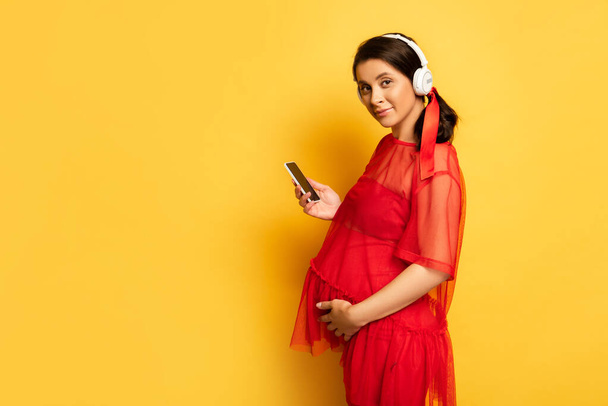 mujer embarazada en auriculares inalámbricos sosteniendo teléfono inteligente mientras toca la barriga en amarillo - Foto, Imagen