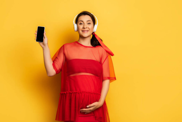 вагітна жінка в бездротових навушниках, що показують смартфон, торкаючись животика на жовтому
 - Фото, зображення