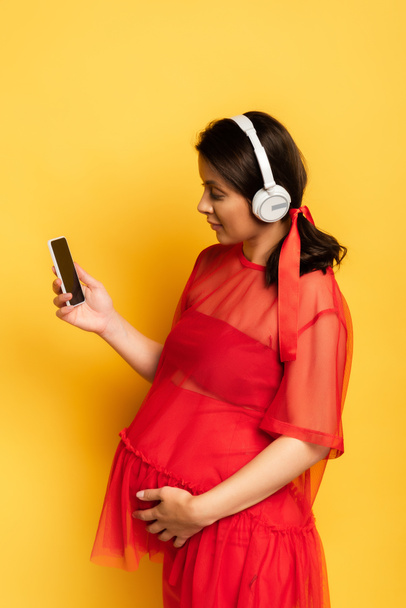 young woman in wireless headphones holding smartphone and touching tummy on yellow - 写真・画像