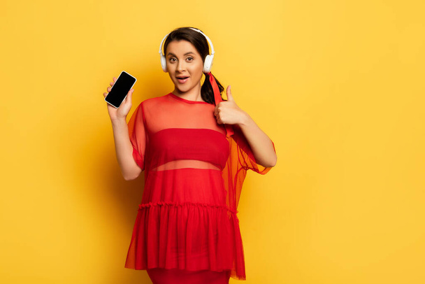 junge Frau in roter Tunika zeigt Smartphone mit leerem Bildschirm und Okay-Geste auf gelb - Foto, Bild
