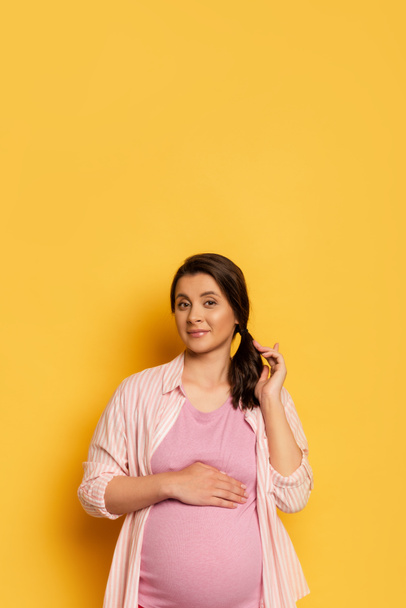mujer embarazada tocando el vientre y el pelo mientras mira a la cámara en amarillo con espacio de copia - Foto, Imagen