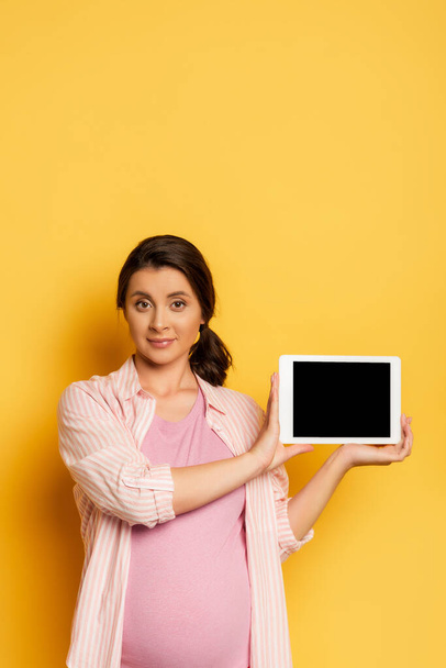 pregnant woman showing digital tablet with blank screen and looking at camera on yellow - Fotoğraf, Görsel