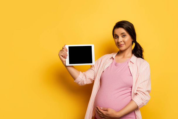 pregnant woman touching belly while showing digital tablet with blank screen on yellow - Zdjęcie, obraz
