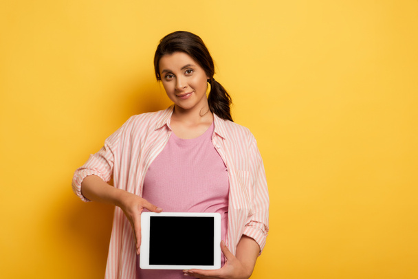 young pregnant woman showing digital tablet with blank screen while looking at camera on yellow - Foto, imagen