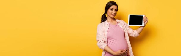 horizontal image of young pregnant woman showing digital tablet with blank screen on yellow - Fotó, kép