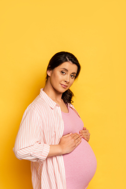νεαρή έγκυος γυναίκα αγγίζει την κοιλιά, ενώ κοιτάζοντας κάμερα στο κίτρινο - Φωτογραφία, εικόνα