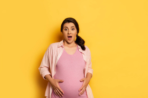 shocked pregnant woman looking at camera while touching belly on yellow - 写真・画像