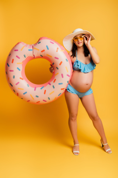 mujer embarazada joven tocando gafas de sol mientras sostiene el anillo de natación en amarillo - Foto, imagen