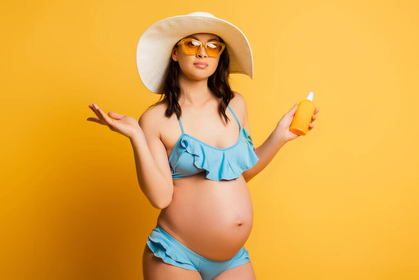 έγκυος γυναίκα με μαγιό και ψάθινο καπέλο κρατώντας αντηλιακό στο κίτρινο - Φωτογραφία, εικόνα