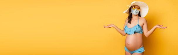 mujer embarazada confundida en traje de baño y máscara médica que muestra gesto de encogimiento de hombros en amarillo - Foto, Imagen