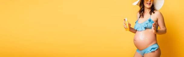 vista ritagliata della donna incinta che applica la protezione solare sulla pancia mentre posa su giallo, colpo panoramico - Foto, immagini