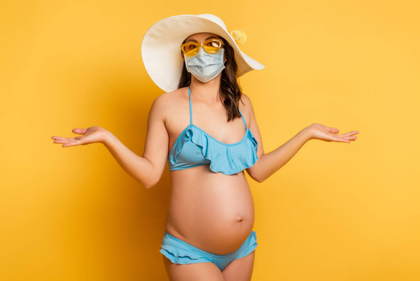 verward zwangere vrouw in medisch masker en badpak tonen schouderophalen gebaar op geel - Foto, afbeelding