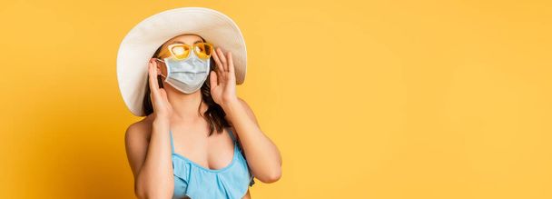 tiro panorámico de la mujer en sombrero de sraw tocando gafas de sol y máscara médica en amarillo - Foto, Imagen