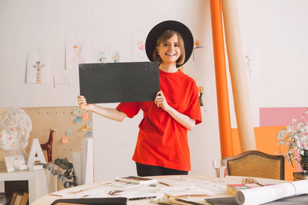 Young cheerful girl with black cardboard sign in hat stand in art studio - Фото, зображення