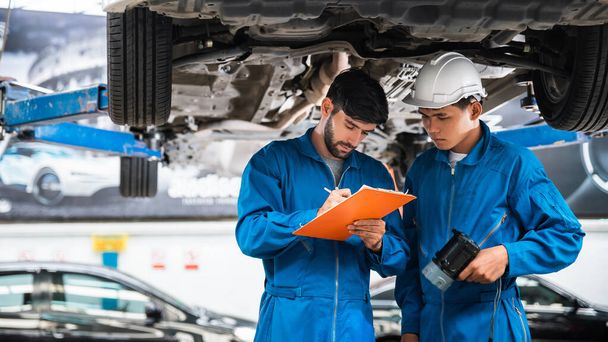 Mechanic in blue workwear uniform checks the vehicle maintenance checklist with his assistant under the lifted car. Automobile repairing service, Professional occupation teamwork. Vehicle maintenance. - Photo, Image
