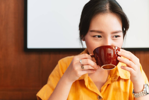 Μια όμορφη Ασιάτισσα που φοράει κίτρινο πουκάμισο πίνει καφέ στο καφέ με χαρούμενα μάτια. Απολαμβάνω τον γυναικείο τρόπο ζωής. Συγκεντρώσου σε ένα φλιτζάνι καφέ.. - Φωτογραφία, εικόνα