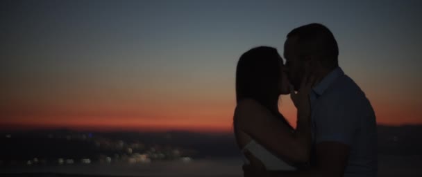 Silhouette di una coppia che si bacia al tramonto con luci notturne sullo sfondo. Rallentatore, DOF superficiale. - Filmati, video
