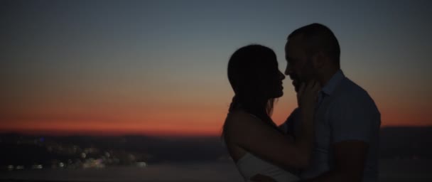 Silhouette di una coppia che si abbraccia al tramonto con luci notturne sullo sfondo. Rallentatore, DOF superficiale. - Filmati, video