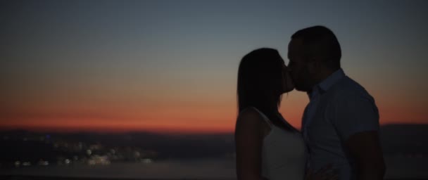 Siluetti pariskunnasta suutelemassa auringonlaskun aikaan yökaupungin valot taustalla. Hidasta, pinnallinen DOF. - Materiaali, video