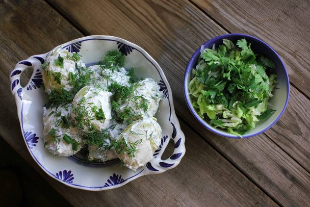 A plate of boiled new potatoes with dill, chopped onion, sour cream, and fresh cabbage salad on the old wooden board table. Top view. Home-cooked healthy food concept - Photo, Image