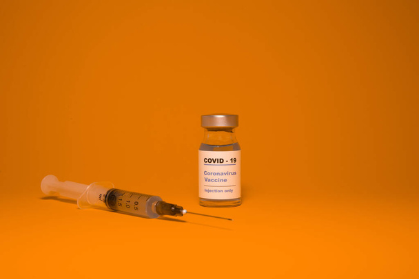 Üzerinde "Covid - 19 Corona virüsü aşısı enjeksiyonu" yazan bir etiket bulunan aşı şişesi (şişe) ve önleme, aşılama ve virüse karşı tedavi için turuncu aşıya izole edilmiş tıbbi bir şırınga - Fotoğraf, Görsel