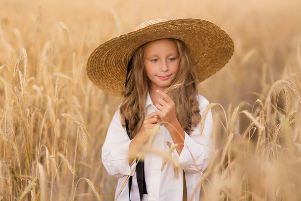Adorable little girl in a straw hat in wheat field. Child with long blonde wavy hair on countryside landscape with spikelet in hand. Farming agriculture harvesting concept. - 写真・画像
