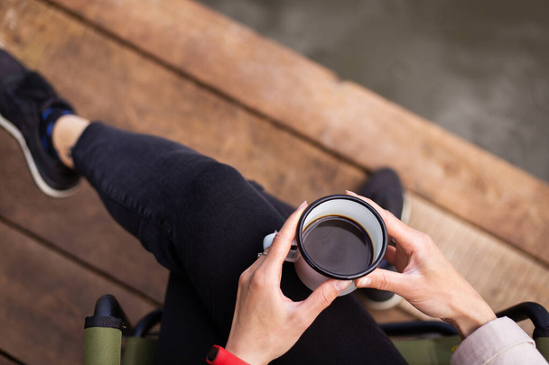 Dziewczyna trzyma metalową filiżankę kawy w dłoniach w pobliżu jeziora, siedząc na drewnianym pomoście. Zbliżenie, widok z góry - Zdjęcie, obraz