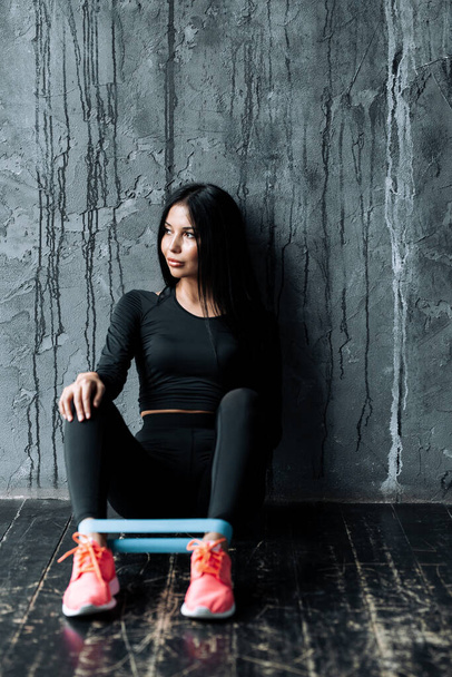 Young woman of pilates trainer in black, tight-fitting clothing, sits on the floor in a loft with a sports rubber expander on her feet. - Фото, зображення