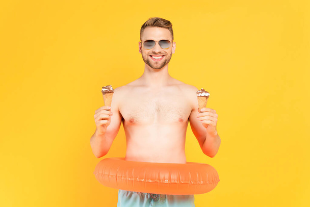 cheerful and shirtless man in sunglasses with inflatable ring holding ice cream cones isolated on yellow - Φωτογραφία, εικόνα