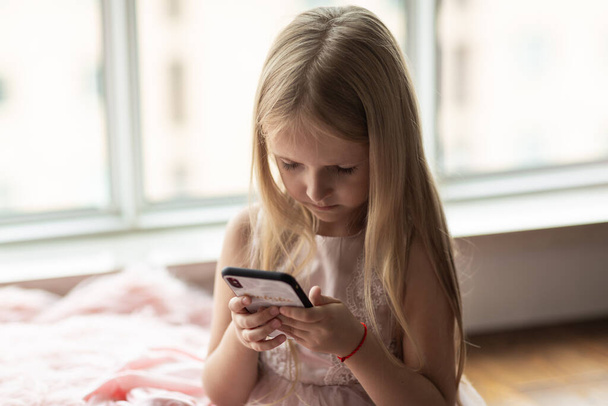 Cute little caucasian girl with blonde hair in fashionable dress sitting at home during coronavirus pandemic quarantine and using mobile phone. Stay at home during covid-19 lockdown concept. High quality photo - Fotoğraf, Görsel