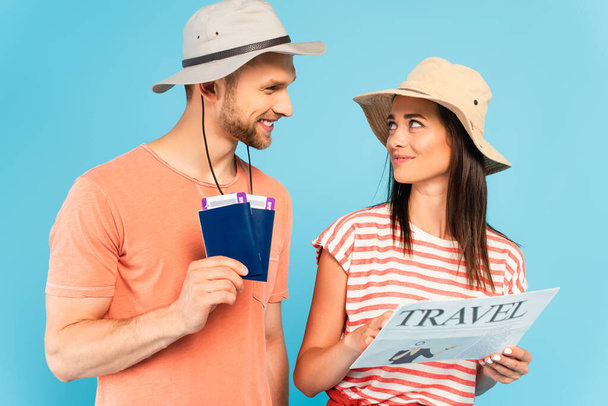 Ευτυχισμένο κορίτσι με καπέλο που κρατάει ταξιδιωτική εφημερίδα και κοιτάζει έναν άντρα με διαβατήρια που είναι απομονωμένος στο μπλε. - Φωτογραφία, εικόνα