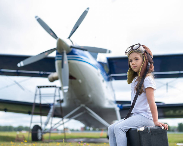 A little girl in a pilots costume sits on a retro suitcase at the airport waiting for the departure of the flight. A child in a hat and glasses is going on a trip by plane. - 写真・画像