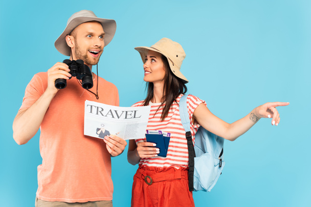 mujer feliz señalando con el dedo y sosteniendo pasaportes cerca de hombre excitado con periódico de viaje y prismáticos aislados en azul  - Foto, imagen
