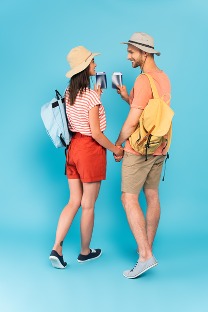 happy couple in hats holding hands and passports while looking at each other on blue - Photo, Image