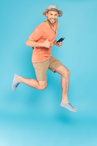 hombre alegre en sombrero con pasaporte, saltando y mostrando el pulgar hacia arriba en azul - Foto, Imagen
