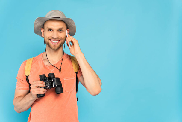 happy man holding binoculars and talking on smartphone isolated on blue - Foto, afbeelding