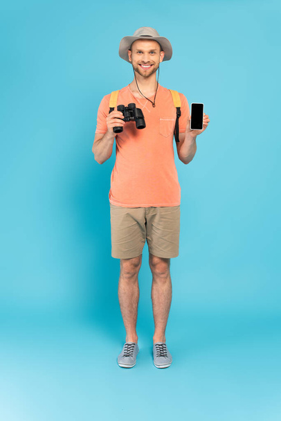 happy man holding binoculars and smartphone with blank screen on blue - Foto, Imagem