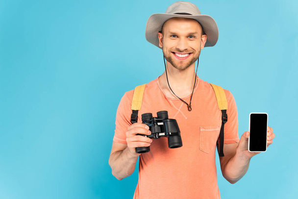 cheerful man holding binoculars and smartphone with blank screen on blue - Fotoğraf, Görsel