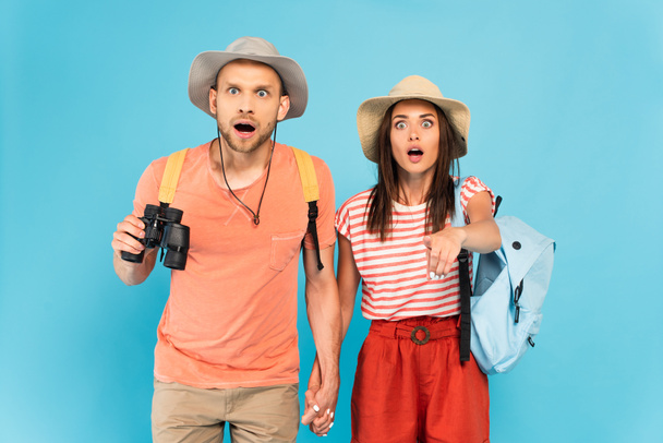 shocked woman in hat pointing with finger near boyfriend with binoculars isolated on blue  - Photo, Image
