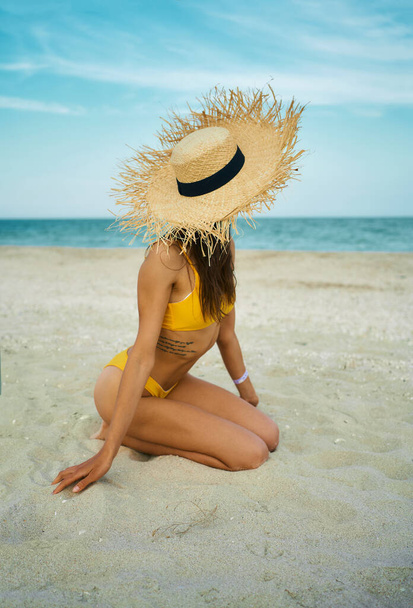 vertical shot sexy bikini tanned body woman wearing big straw hat sitting on sand beach by blue sea. - Photo, image