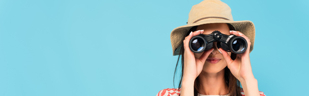 panoramic shot of young woman in hat looking through binoculars isolated on blue - Foto, imagen