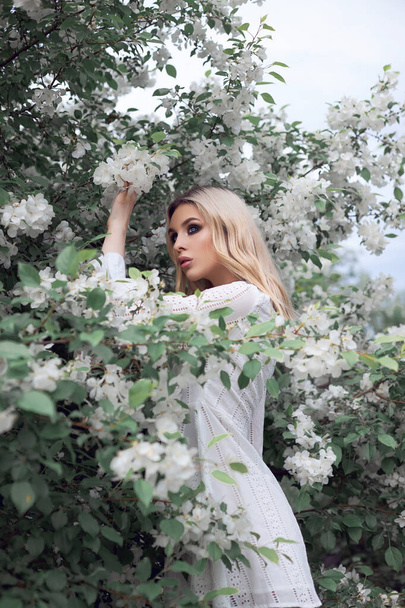 Sexy blonde woman in summer in the branches of an Apple tree. Portrait of a girl in white Apple blossoms - Foto, afbeelding