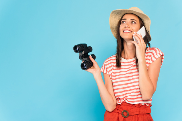 happy woman in hat talking on smartphone and holding binoculars on blue - Fotó, kép